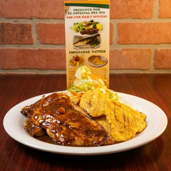 Plate of sauced meat, fried plantains, and salad on a table with a daily special menu.